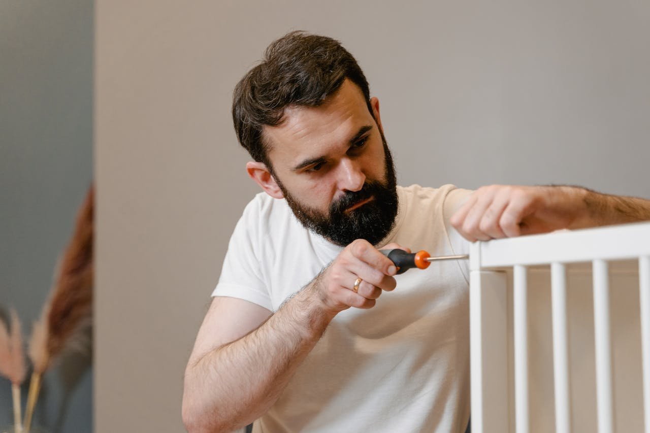 A man focusing closely on assembling a baby crib with a screwdriver in a well-lit room.